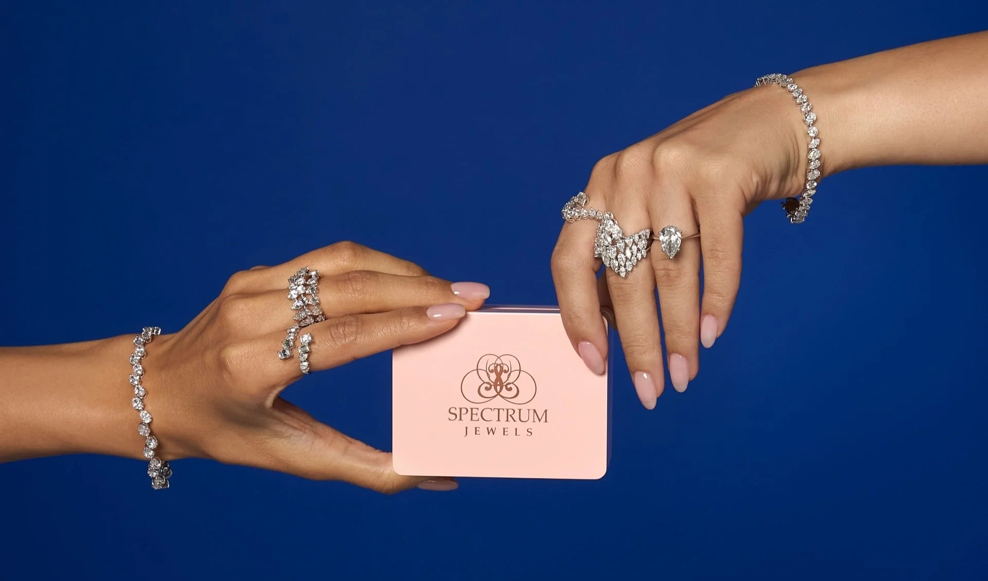 Spectrum Jewels hands holding a pink jewelry box, showcasing diamond rings, tennis bracelets, and fine diamond jewelry against a blue background.