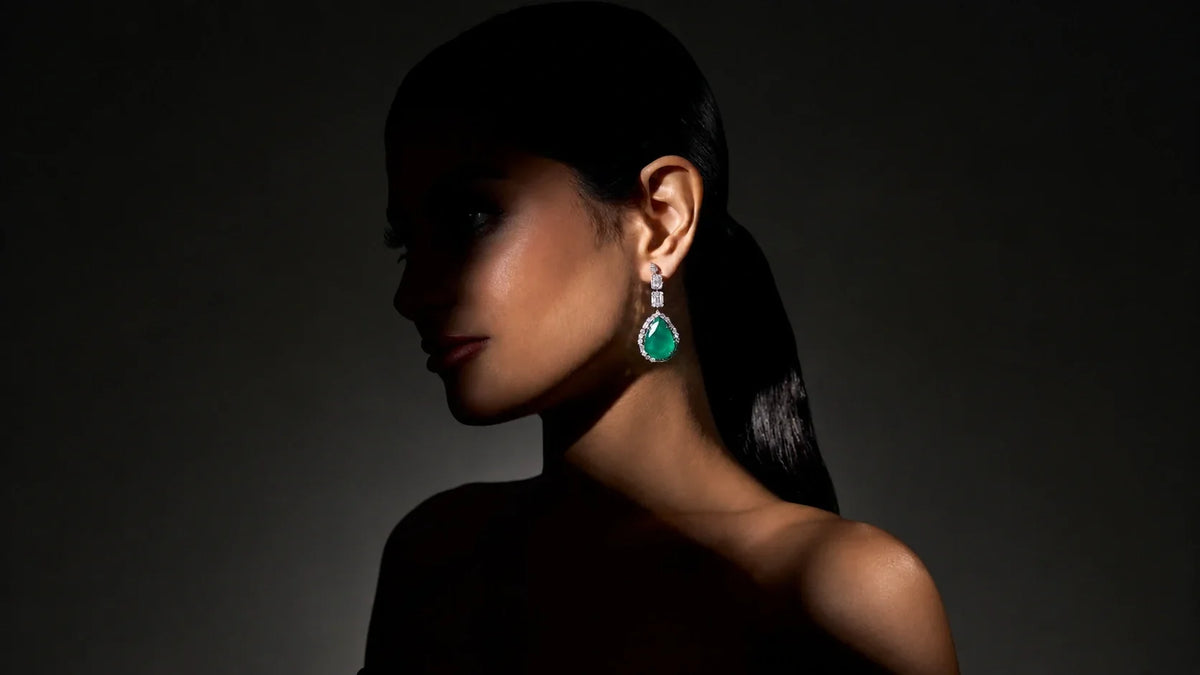 A model wearing Spectrum Jewels' pear-shaped emerald and diamond drop earring against a dark evening backdrop.