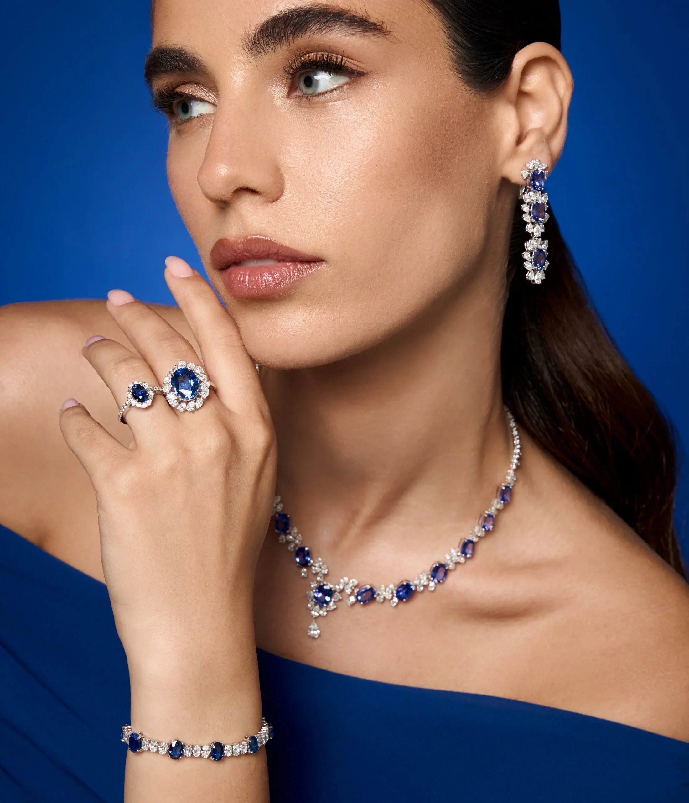 Spectrum Jewels model wearing a blue sapphire and diamond jewelry set featuring drop earrings, a gemstone necklace, a bracelet, and matching sapphire rings in white gold.