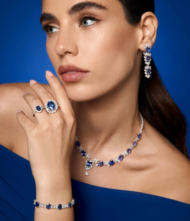 Spectrum Jewels model wearing a blue sapphire and diamond jewelry set featuring drop earrings, a gemstone necklace, a bracelet, and matching sapphire rings in white gold.